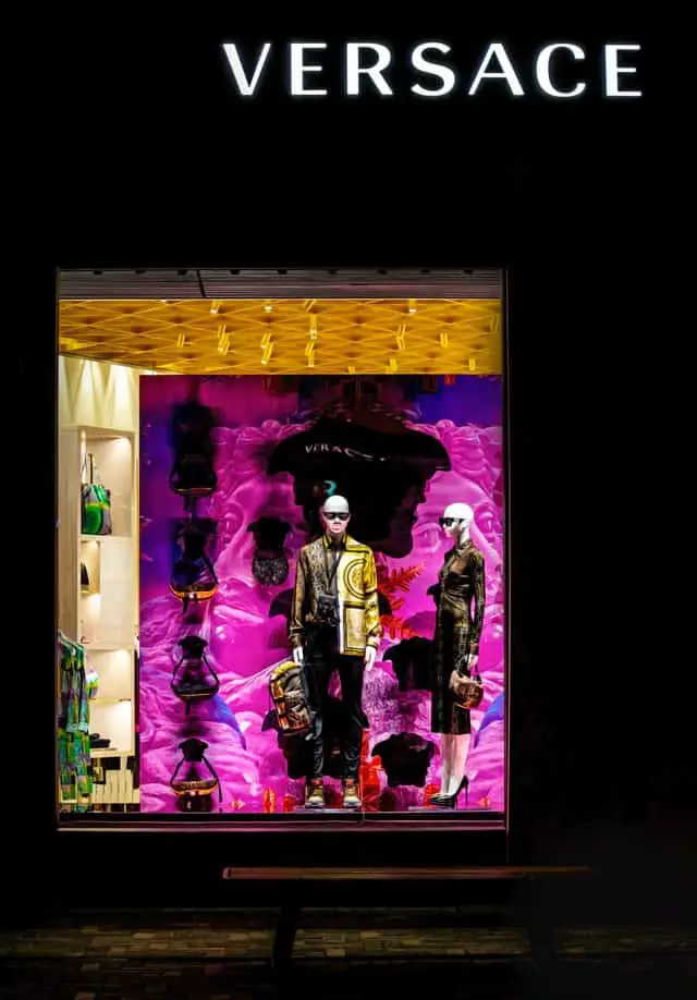 A Versace store window with a black background and white letters, featuring a colorful display with both patterns and a male and female mannequin.
