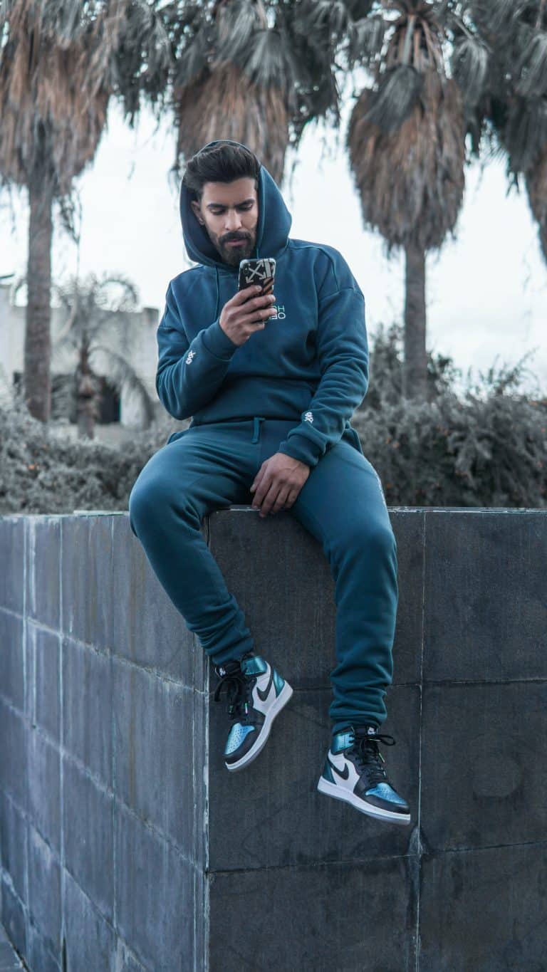 Man sitting on a grey wall wearing a blue tracksuit, and blue, white and black Nike shoes. He is holding a phone with an OFF-WHITE phone case against a cloudy background, with some palm trees.