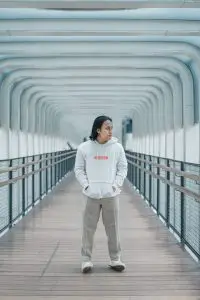 Man with long, dark hair stands in a tunnel with a white hoodie, khaki pants, and white sneakers.
