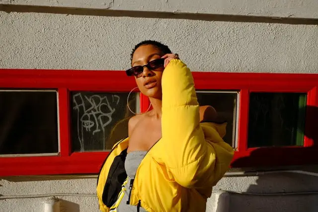 Black woman with natural short hair in gold hoop earrings, sunglasses, yellow puffer jacket, and grey dress looks at the camera in front of red windows.