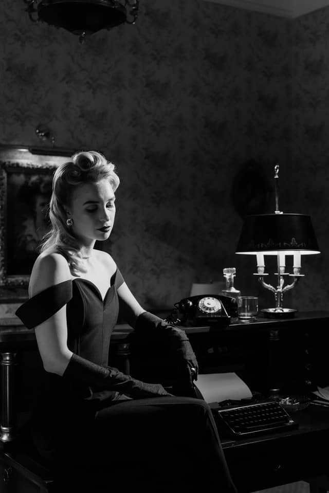 Black and white photograph of a blonde woman in a dark room with a black dress and long black gloves