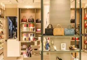 Department store window with black, yellow, and blue handbags in the foreground and luxury shoes on the shelves.