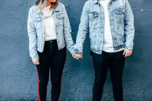 Man and Woman holding hands wearing light acid washed denim jackets against a yale blue background