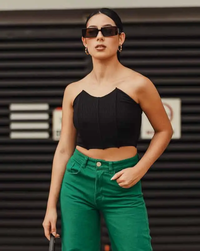 A Brunette girl wearing trendy, rectangular, black sunglasses, a strapless black corset top, bright green jeans, and gold hoop earrings. The girl’s hair is in a slicked back ponytail and she is standing in front of a black background that looks like a garage door.